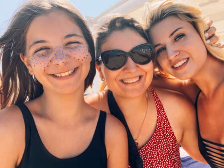 Company of friendly women in swimwear relaxing on beach on sunny day and taking selfie while smiling and looking at cameraの写真素材