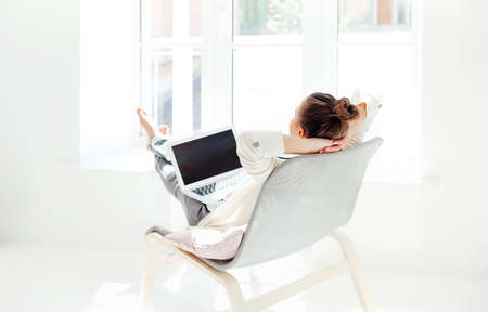 Barefoot young female with hands behind head sitting on chair and relaxing with closed eyes after using laptop in light room at homeの写真素材