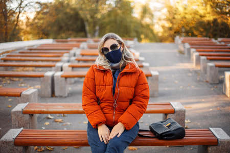 Blond female in stylish outerwear and fabric mask looking at camera while stitting on benche in park during pandemicの写真素材