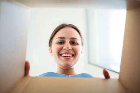 From below happy young female smiling and looking inside carton box while opening ordered goods