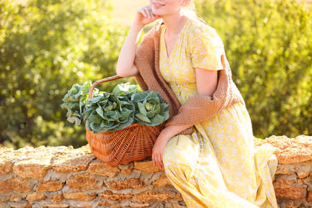 Unrecognizable young female leaning on wicker basket with fresh cabbage and touching face while resting on brick fence on sunny summer day in countrysideの写真素材