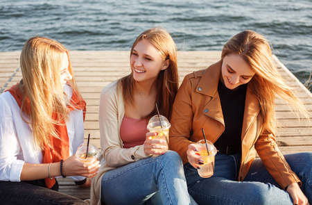 Happy young women with takeaway drinks walking on pavement and smiling on sunny weekend day on city streetの写真素材