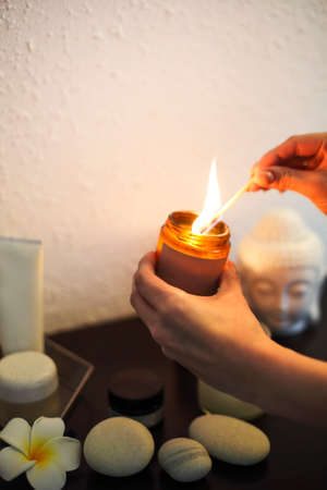 Faceless woman lighting candle in jar with burning chip above table with spa stones