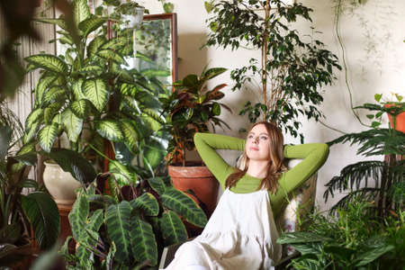 Calm female with hands behind head lounging on chair in room all filled with flowerpots of lush plantsの写真素材