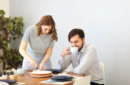 Woman cutting pie and man drinking beverage while having breakfast together at table in kitchen