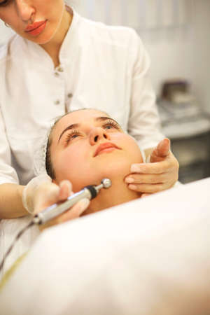 Crop cosmetologist applying modern machine for microcurrent massage on face of woman lying on table...
