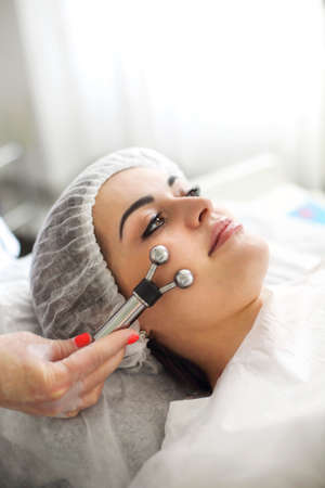 Unrecognizable crop cosmetologist applying modern machine for microcurrent massage on face of woman...
