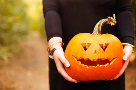 Young woman in black cape carrying spooky jack o lantern and looking away on Halloween day in autumn forestの写真素材