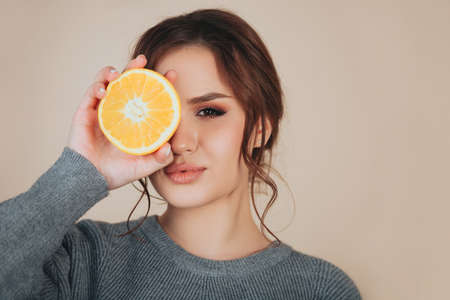 Brunette female wearing wooven gray sweater holding orange slice next to eyeの写真素材