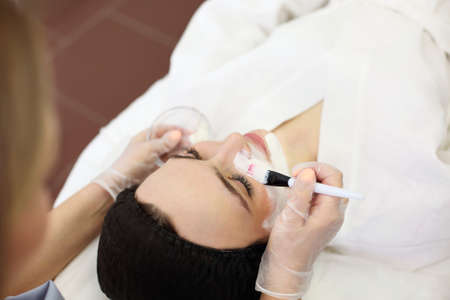 Unrecognizable female beautician applying clay mask with brush on face of relaxed client while doing skincare procedure in spa salonの写真素材
