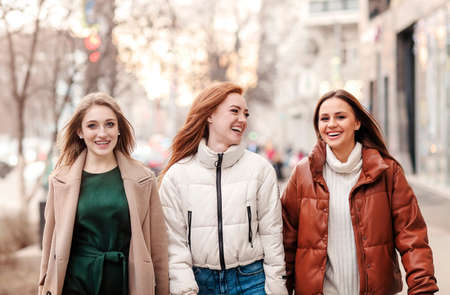 Optimistic girlfriends in outerwear smiling and walking on street of modern city togetherの写真素材