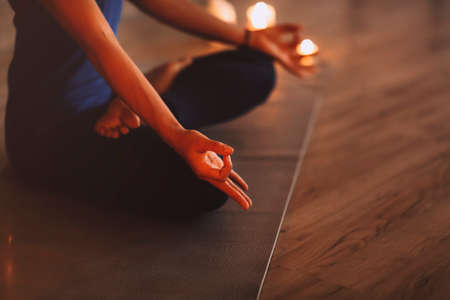 Young female meditating with closed eyes and clasped hands while sitting cross legged near burning candles in evening in cozy room. Close upの写真素材