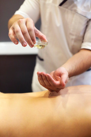 Crop faceless masseuse pouring oil on hand during relaxing massage in spa centerの写真素材