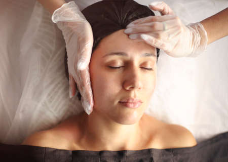 Unrecognizable masseuse in gown standing in spa salon and massaging face of female customer