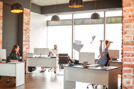 Happy woman throwing papers into air and screaming in excitement near colleagues after finishing project in modern officeの写真素材