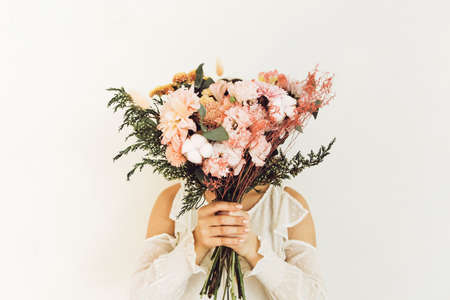 Unrecognizable female covering face with bouquet of fresh flowers against gray background in summerの写真素材