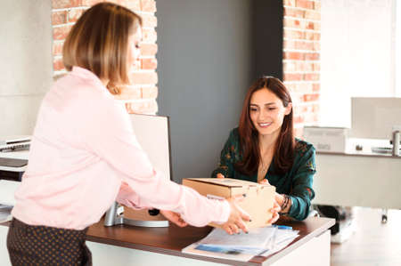 Cheerful female manager smiling and taking carton box from delivery woman while working in officeの写真素材