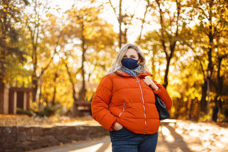 Blond woman in outerwear and mask walking on paved path on sunny day in autumn park during pandemic