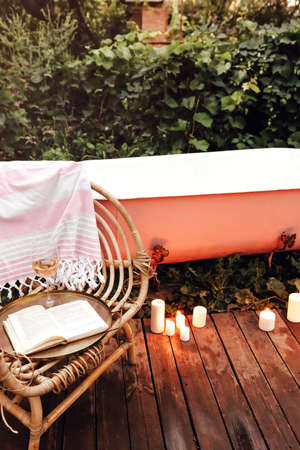 Burning candles and wicker chair with book and wine placed on wooden path near bathtub in garden