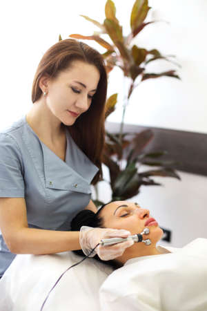 Cosmetologist applying modern machine for microcurrent massage on face of woman lying on table in...