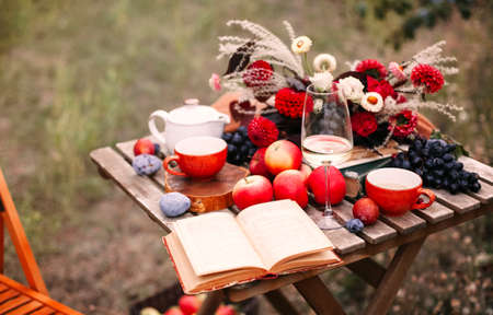 Opened book and glass of wine placed near dishware and bouquet of fresh flowers amidst assorted fruits on wooden table on autumn day in gardenの写真素材