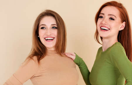 Happy redhead female touching shoulder of girlfriend and looking away with smile against beige backgroundの写真素材