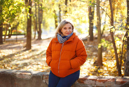 Delighted female in outerwear smiling and covering eye with dry leaf on sunny weekend day in autumn...