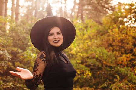 Young woman in black cape carrying spooky jack o lantern and looking away on Halloween day in autumn forestの写真素材