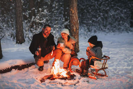 Cheerful parents and son enjoying hot drink while resting near campfire in evening in winter forestの写真素材