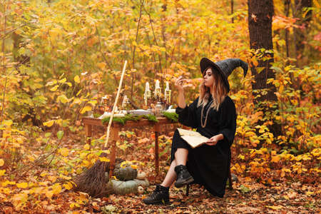 Young woman in witch costume sitting at table with magic ingredients and making notes on autumn day in forestの写真素材