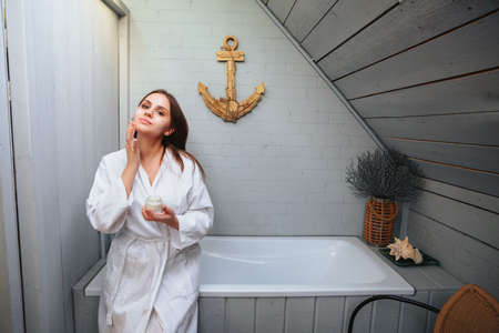 Beautiful brunette female in bathrobe sitting on edge of bathtub in bathroom while gently applying moisturizing cream on face, morning hygiene procedures routine and skincare beauty treatment conceptの写真素材
