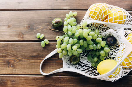 Top view of knitted grocery mesh with organic fruits flat lay on wooden table surface background, bunches of grapes and lemons lie falling from bag. Food store delivery conceptの写真素材