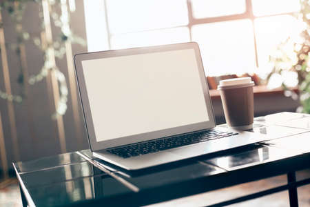 Photo of white blank screen of laptop with keyboard case lying on little table with disposable cup of coffee from cafeteria next to computer over comfortable living room interior backgroundの写真素材