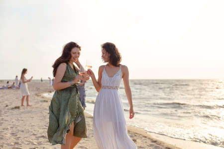 Glad adult female friends in summer outfit smiling while enjoying wine on sandy beach near seaの写真素材