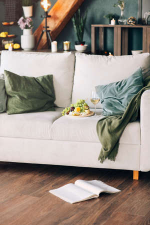 Plate with fresh fruits and glass of wine placed near pillows on comfortable couch in stylish room at homeの写真素材