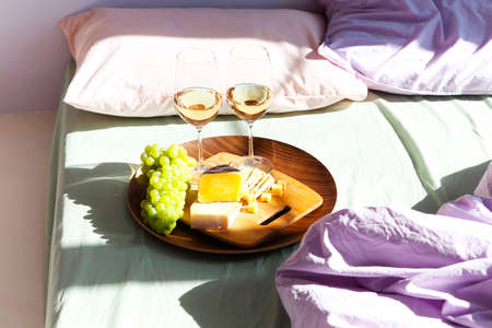 Glasses of red wine and plate with assorted food placed on comfortable bed during romantic date