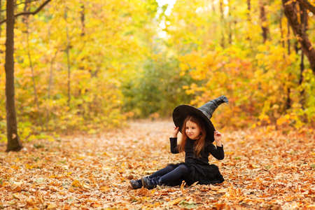 Cute happy little redhaired girl dressed in witch costume sitting on fallen leaves over autumn forest background with copy space, holding her hat with hands. Holiday of halloween celebration conceptの写真素材