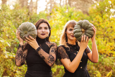 Two female friends holding scary green pumpkins in front of her face, standing isolated over autumn forest background. Fall holidays, halloween decorations and people conceptの写真素材