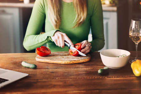Cropped photo of unknown blond woman cutting and slicing tomatoes with sharp knife in kitchen on wooden table top, preparing vegetable salad. Cooking healthy, organic food and homemade conceptの写真素材