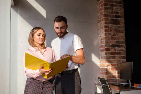 Two young focused coworkers, woman and man, looking through documents from folder standing in office. Business partners discussing financial graphs and reports together. Teamwork conceptの写真素材