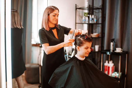 Positive woman hairdresser at working process in beauty salon, professional smiling hairstylist preparing female client for haircut, pinning up hair with clips. Business, fashion and beauty conceptの写真素材
