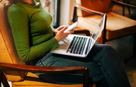 Side view of happy woman smiling and browsing laptop while sitting at table and working on remote project in cafeteriaの写真素材