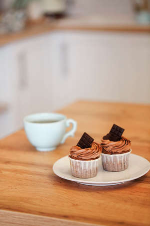 Close-up image of two muffins on white plate decorated with cocoa cream and pieces of chocolate with cup of tea on background, served on wooden table in coffee shop. Pastries and dessert conceptの写真素材