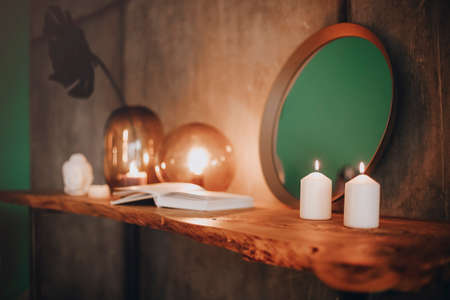 Round mirror on wooden shelf with two burning candles and small porcelain Buddha head reflected in corner of mirrored surface over concrete wall background in minimalist modern apartment interiorの写真素材