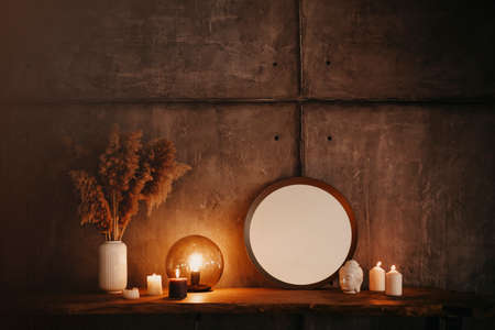 Round mirror on wooden shelf with two burning candles and small porcelain Buddha head reflected in corner of mirrored surface over concrete wall background in minimalist modern apartment interiorの写真素材