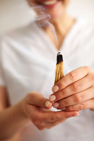 Atmospheric photo of smiling female SPA worker in white uniform holding burning palo santo incense stick with smoke slowly rising up. Spiritual practice, energy cleanse, healing, meditation conceptの写真素材