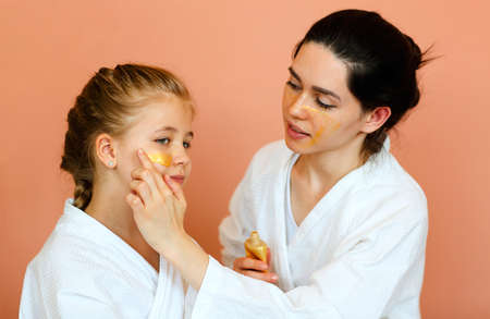 Family spa day. Young beautiful mother and little daughter in bathrobes doing beauty treatment together, mom applying gold colored face mask on child face, standing isolated over orange backgroundの写真素材