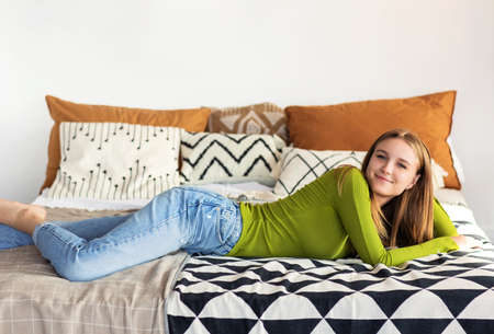 Smiling positive teen girl in casual clothes lying on bed in her cozy room at home, resting after...