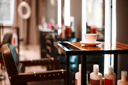 Hairdressing salon interior. Coffee cup on wooden table and chair for client in beauty parlor with nobody, illuminated mirror in blurred background, empty hairdresser workplaceの写真素材