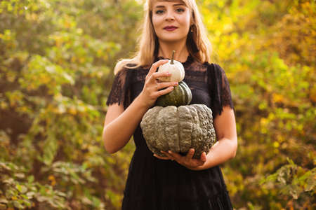 Portrait of beautiful young blond woman with long straight hair and dark vamp makeup looking to the camera, wearing black guipure dress, holding pumpkins while standing against autumn forest backgroundの写真素材